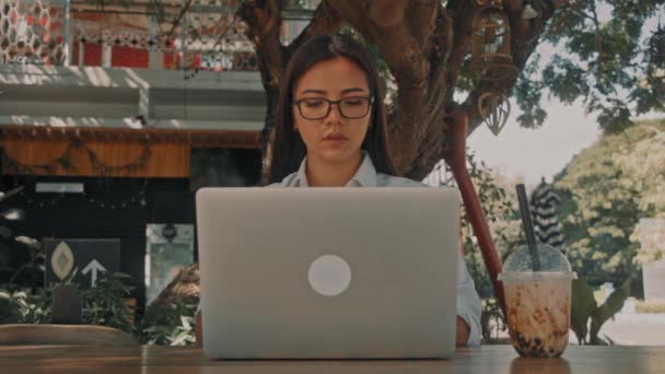 Asiático atraente jovem mulher vestindo óculos, trabalhando em seu laptop em um café terraço ao ar livre — Vídeo de Stock