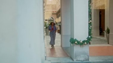 Güzel, modaya uygun Afro-Amerikan bir kadın Noel süslerinin önünde sokakta yürüyor.