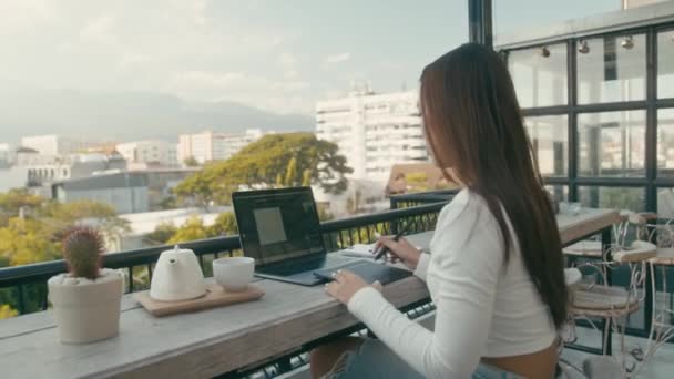 Egy fiatal ázsiai lány dolgozik a laptopjánál egy háztetőn a városban. — Stock videók