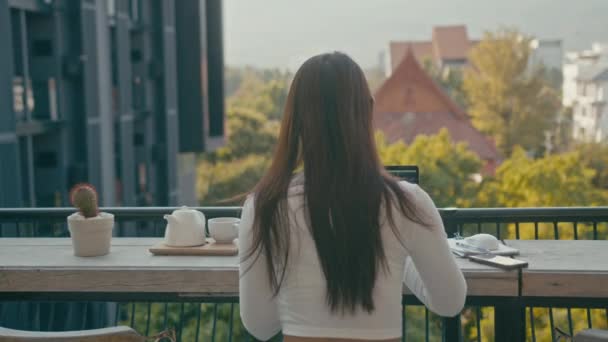 Una joven asiática trabaja en su portátil en un café en la azotea de la ciudad — Vídeos de Stock
