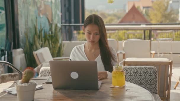 アジア系の少女がノートパソコンで街の屋上カフェで働いています — ストック動画