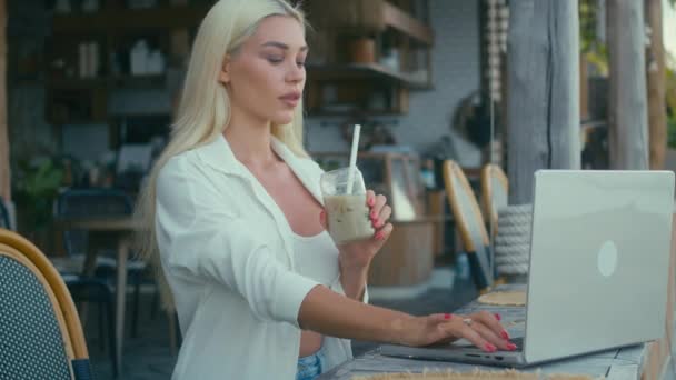白いシャツを着たブロンドの女の子は カフェでラップトップで働いています カメラを見て フルーツシェイクを飲みます フリーランスのコンセプト — ストック動画