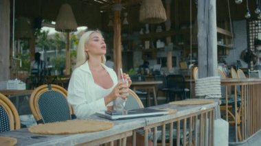 Beyaz gömlekli sarışın kız kafede dizüstü bilgisayarla çalışıyor, kameraya bakıyor ve meyve suyu içiyor. Bağımsız kavram