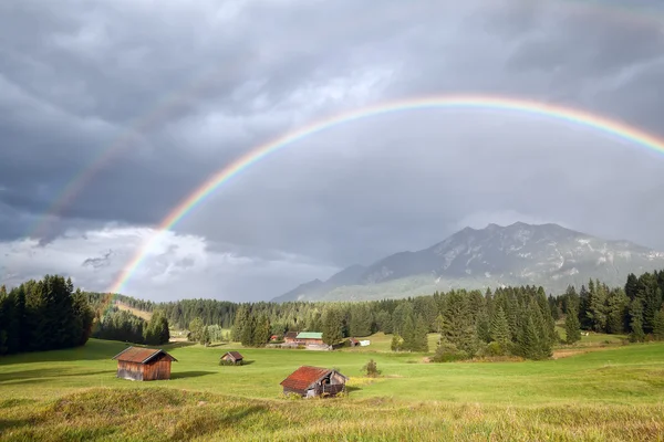 Красочная радуга над альпийскими лугами с деревянными хижинами — стоковое фото