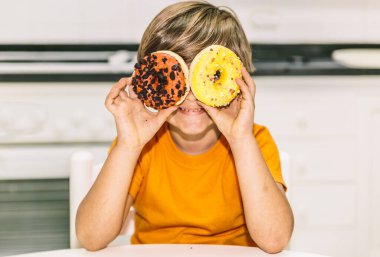 Sevimli çocuk evde mutfakta gülümserken iki donuta bakıyor.