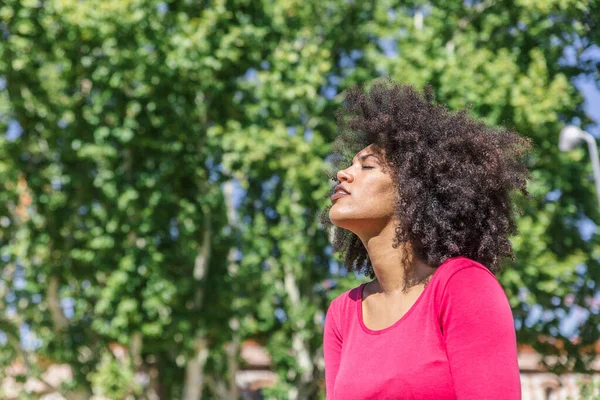 Afro saçlı çekici bir kadın ağaçlarla çevrili bir parkta rahatça nefes alıyor.