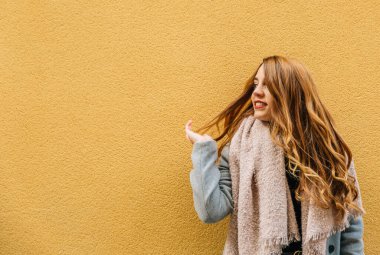 Sarışın kız, sarı bir duvarın önünde saçlarına dokunurken gülümsüyor.