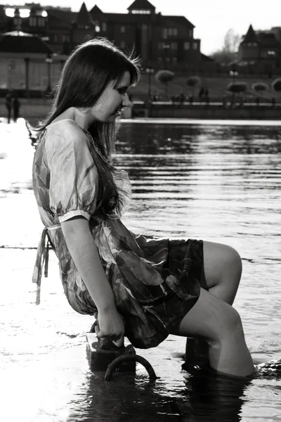 Jovem mulher sentada no aterro e segurando as pernas na água — Fotografia de Stock