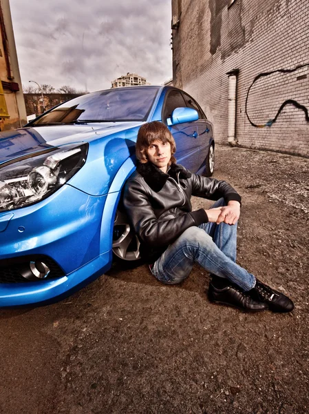 Homme en manteau de cuir assis penché sur la roue avant de la voiture bleue — Photo