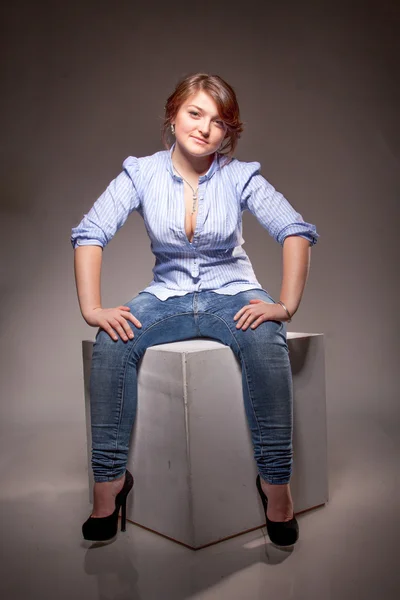 穿着蓝色衬衫和牛仔裤坐在演播室的白色立方体上的女人 — 图库照片