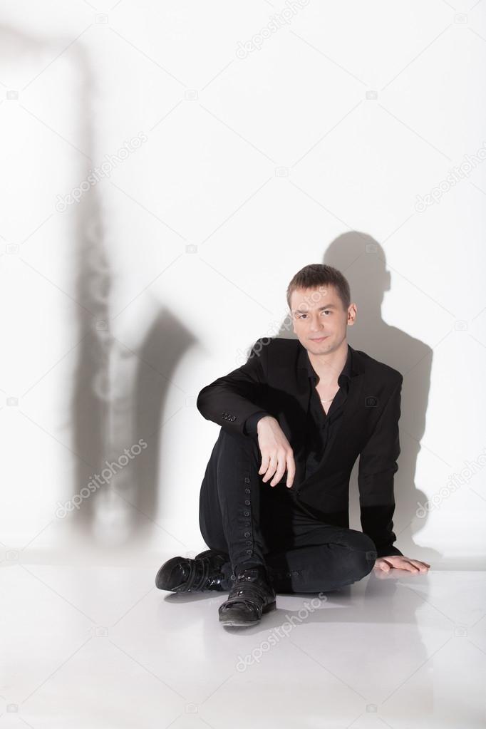 Man in black suit sitting on floor against shadow of saxophone