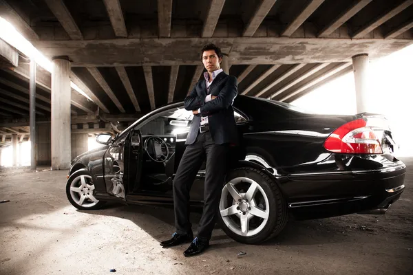 Man in suit leaning against black expensive car under bridge — Stock Photo, Image