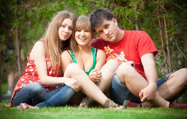 Dos chicas y un joven sentado en el prado con las piernas cruzadas —  Fotos de Stock