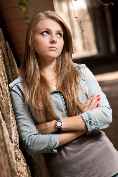 Blond meisje in jeans vacht leunend tegen muur en opzij op zoek — Stockfoto