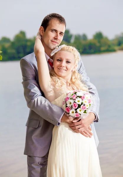 Tall groom hugging blonde bride from back against river — Stock Photo, Image