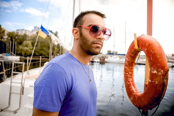 Man in sunglasses standing at seaport against yachts and lifebuoy — Stock Photo, Image