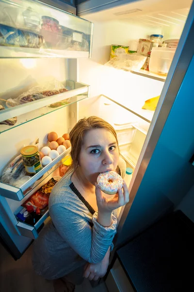 Mulher de pijama comendo donut ao lado da geladeira — Fotografia de Stock