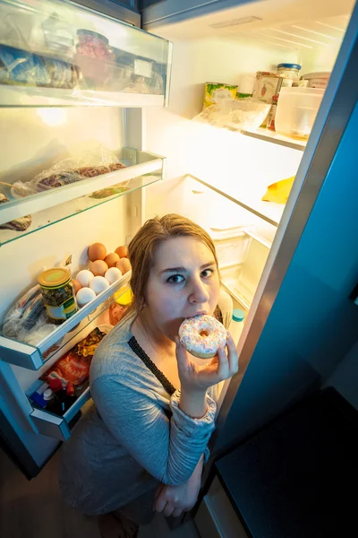 Frau im Pyjama isst Donut neben Kühlschrank — Stockfoto