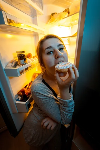 Retrato de la mujer mordiendo donut en la noche cerca de la nevera — Foto de Stock