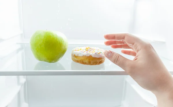 Hand choosing between apple and donut at fridge — Stock Photo, Image