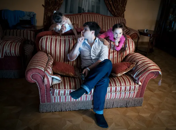 Dos hijas molestando padre mientras mira la televisión por la noche — Foto de Stock