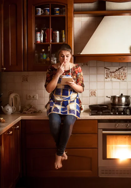 Housewife sitting on tabletop and looking for recipe in tablet — Stock Photo, Image