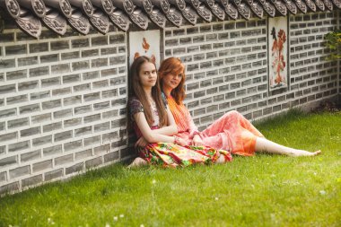 Two girls sitting at ancient korean park against brick wall clipart