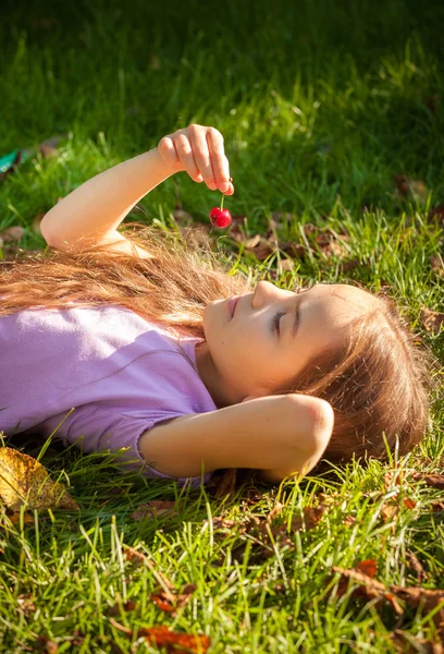 Маленькая девочка лежит на траве и смотрит на красную вишню — стоковое фото