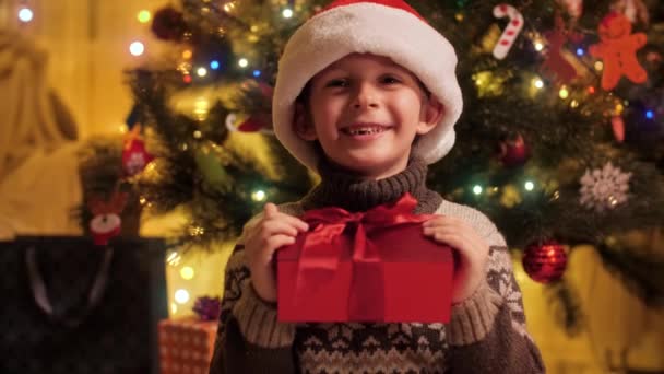 Retrato de niño sonriente alegre con caja de regalo de Navidad con sombrero de Santa sentado al lado del brillante árbol de Navidad decorado. Familias y niños celebrando las vacaciones de invierno. — Vídeo de stock