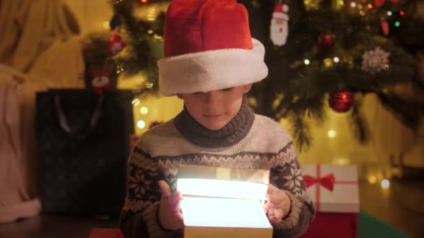 Портрет взволнованного мальчика в шляпе Санты открывает коробку с подарками на Рождество и смотрит внутрь. Волшебство зимних праздников. Семьи и дети празднуют зимние праздники. — стоковое видео