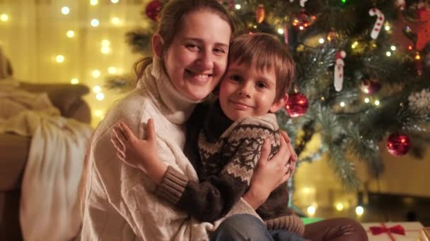 Vidám mosolygós fiú portréja, anya ölelkezik a karácsonyfa mellett, és kamerába néz. A téli ünnepeket ünneplő családok és gyermekek. — Stock videók