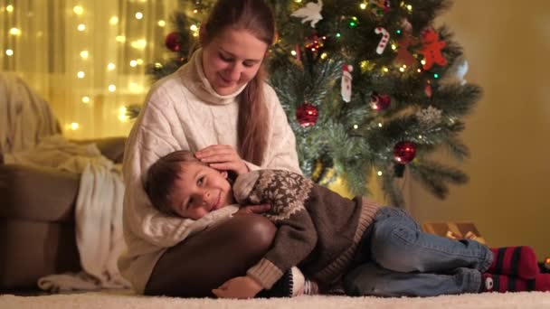 Gelukkig lachende jongen liggend op moeders schoot terwijl ze hem streelde naast de kerstboom. Gezinnen en kinderen vieren wintervakantie. — Stockvideo