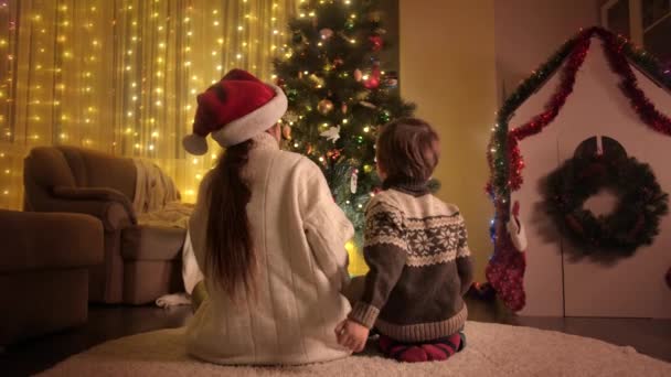 Vista trasera del niño y la madre abrazándose bajo el árbol de Navidad en la sala de estar. Familias y niños celebrando las vacaciones de invierno. — Vídeos de Stock