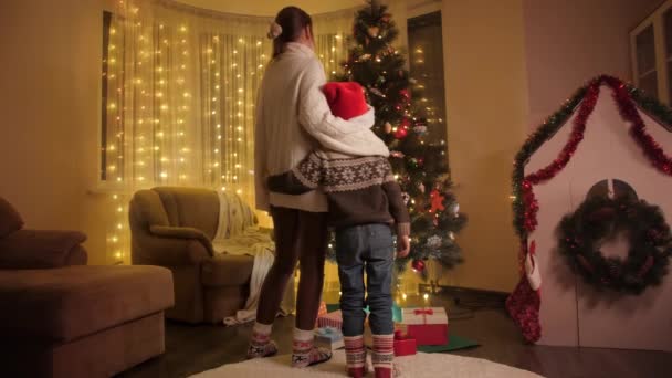 Anya fia a Mikulás kalap áll a nappaliban, és nézi a színes izzó karácsonyfa. A téli ünnepeket ünneplő családok és gyermekek tiszta érzelmei. — Stock videók