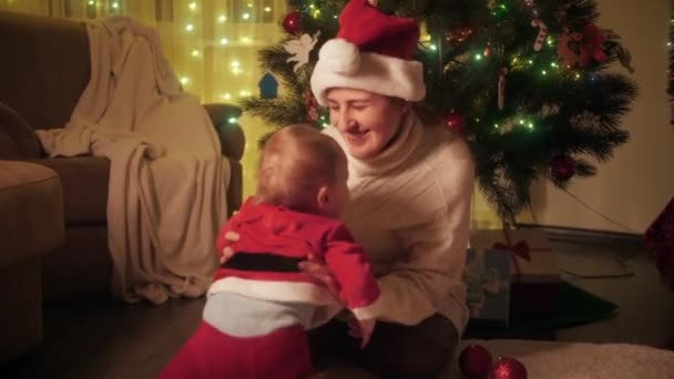 Portret van een vrolijke lachende moeder die thuis met een schattig zoontje onder de kerstboom speelt. Gezinnen en kinderen vieren wintervakantie. — Stockvideo