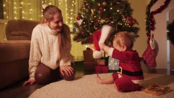 Söt liten pojke klädd i tomte kostym sitter med mamma bredvid julgran. Familjer och barn som firar vinterlov. — Stockvideo