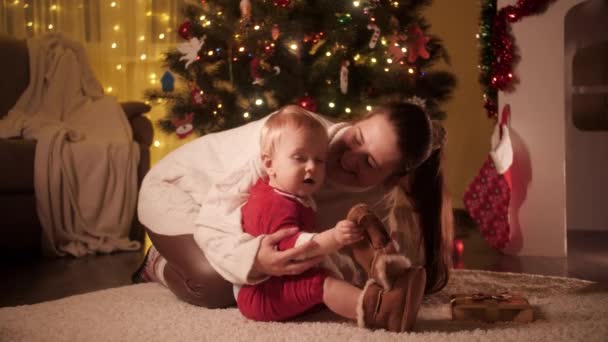 Joven madre sonriente vistiendo bota caliente a su hijo pequeño en traje de Papá Noel al lado del árbol de Navidad. Familias y niños celebrando las vacaciones de invierno. — Vídeos de Stock