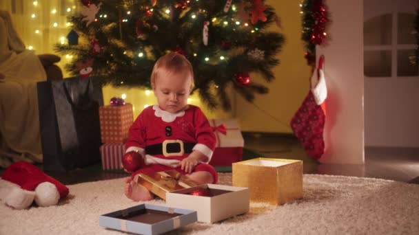 Щасливий усміхнений веселий хлопчик відкриває різдвяні подарунки і повзає на підлозі під ялинкою у вітальні. Сім'ї та діти святкують зимові свята . — стокове відео