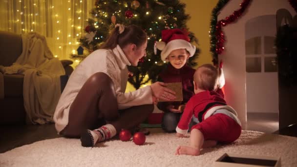 Glückliche Mutter mit Baby und älterem Sohn präsentiert Geschenkboxen neben dem Weihnachtsbaum im Haus. Familien und Kinder feiern Winterurlaub. — Stockvideo