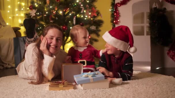 Glücklich lächelnde Kinder, deren Mutter lächelnd neben dem Weihnachtsbaum sitzt und Spaß hat. Familien und Kinder feiern Winterurlaub. — Stockvideo