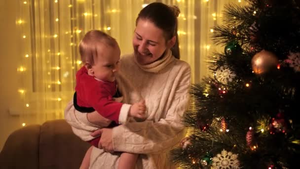 행복하게 미소짓는 엄마가 어린 아들을 안고 빛나는 크리스마스 트리를 바라보고 있습니다. 겨울철 휴일을 기념하는 가족 과 자녀들. — 비디오