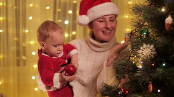Madre sonriente abrazando a su hijo pequeño jugando con adornos y adornos de árboles de Navidad. Familias y niños celebrando las vacaciones de invierno. — Vídeos de Stock