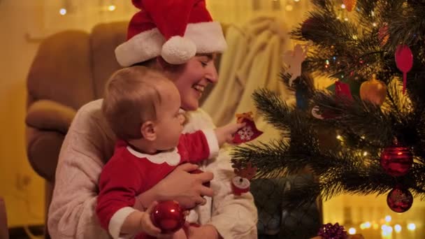 Menino pequeno que espreita na árvore de Natal brilhante e decolando bugigangas de Natal. Famílias e crianças comemorando as férias de inverno. — Vídeo de Stock