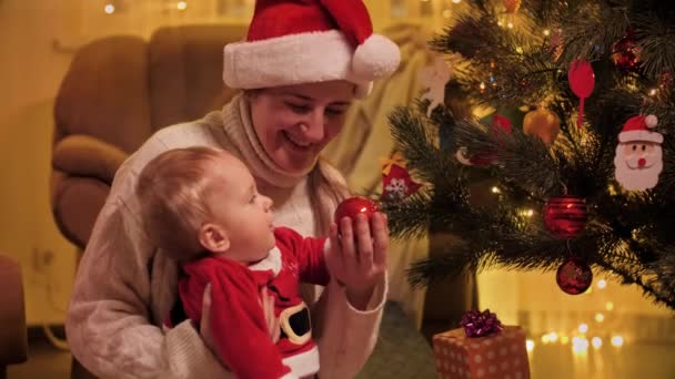 Niño pequeño con madre en el sombrero de Papá Noel decorando el árbol de Navidad con chucherías. Familias y niños celebrando las vacaciones de invierno. — Vídeos de Stock