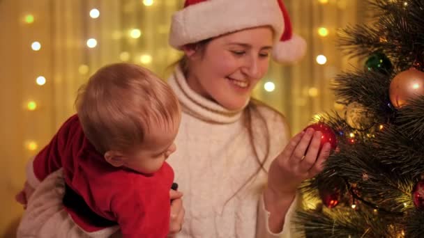小さな赤ちゃんの息子にクリスマスツリーを示す幸せな笑顔の母親。冬の休日を祝う家族や子供. — ストック動画