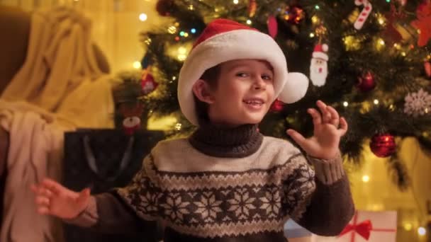 Feliz niño sonriente con sombrero de Santa y agitando la cabeza bajo el árbol de Navidad en la sala de estar. Familias y niños celebrando las vacaciones de invierno. — Vídeos de Stock