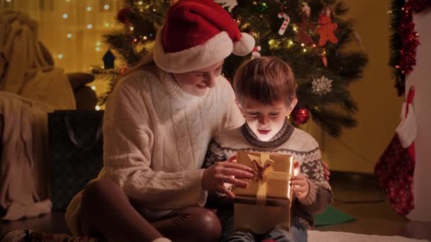 Feliz madre abrazando a su hijo desenvolviendo regalos de Navidad y presentes en la sala de estar. Familias y niños celebrando las vacaciones de invierno. — Vídeos de Stock