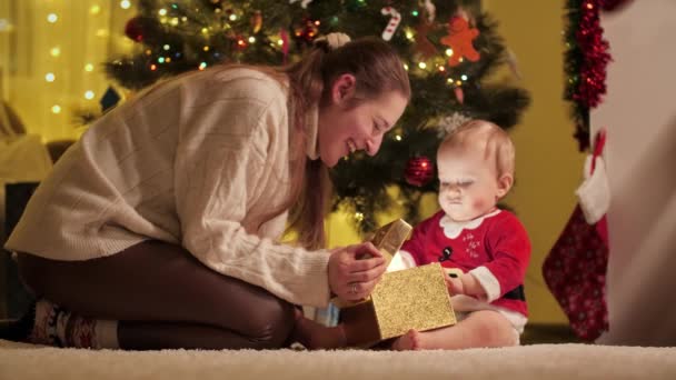 Madre feliz con hijo bebé desenvolviendo y mirando dentro de la caja de regalo de Navidad en la sala de estar. Familias y niños celebrando las vacaciones de invierno. — Vídeos de Stock