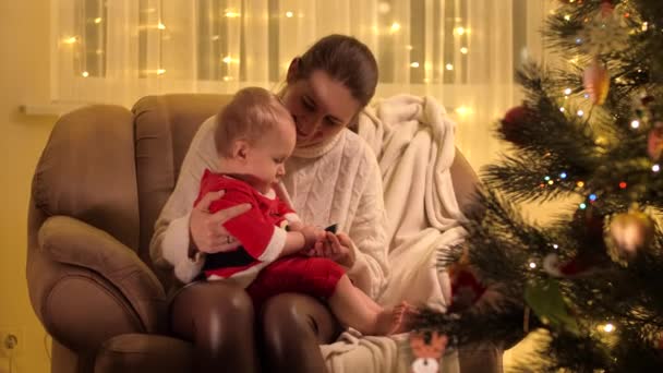 Gelukkig lachende moeder zittend in fauteuil met haar zoontje naast gloeiende kerstboom. Gezinnen en kinderen vieren wintervakantie. — Stockvideo