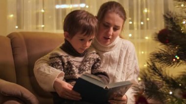 Genç anne oğluna kitap okuyor. Parlayan Noel ağacının yanındaki koltukta oturuyor. Aileler ve çocuklar kış tatilini kutluyor.
